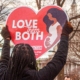 A woman holds a heart-shaped sign that says "Love them both"