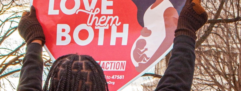A woman holds a heart-shaped sign that says "Love them both"