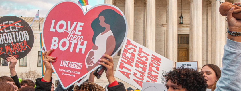 Pro-life people hold signs outside the Supreme Court. One sign says "Love them Both" and another says "Atheist Against Abortion."